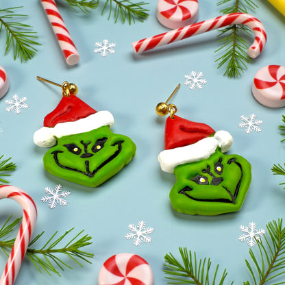 Grinch earrings on a Christmas-themed background with candy canes and pine branches.