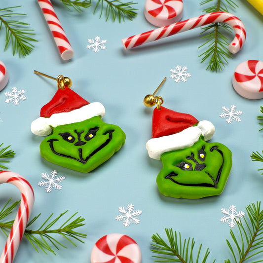 Grinch earrings on a Christmas-themed background with candy canes and pine branches.