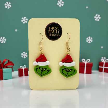 Grinch earrings on a These Pretty Things presentation card with Christmas themed background.