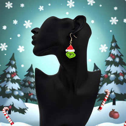 Grinch earrings on a black mannequin head  against a festive themed background with trees and snowflakes.