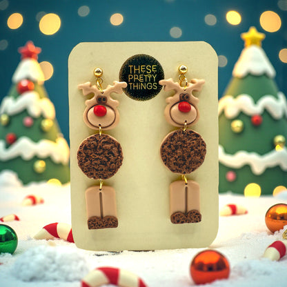 Reindeer earrings on a presentation card with Christmas trees and decorations in the background.