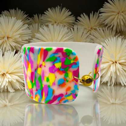 These Pretty Things Confetti Cuff on a table with a cream feather boa in the background
