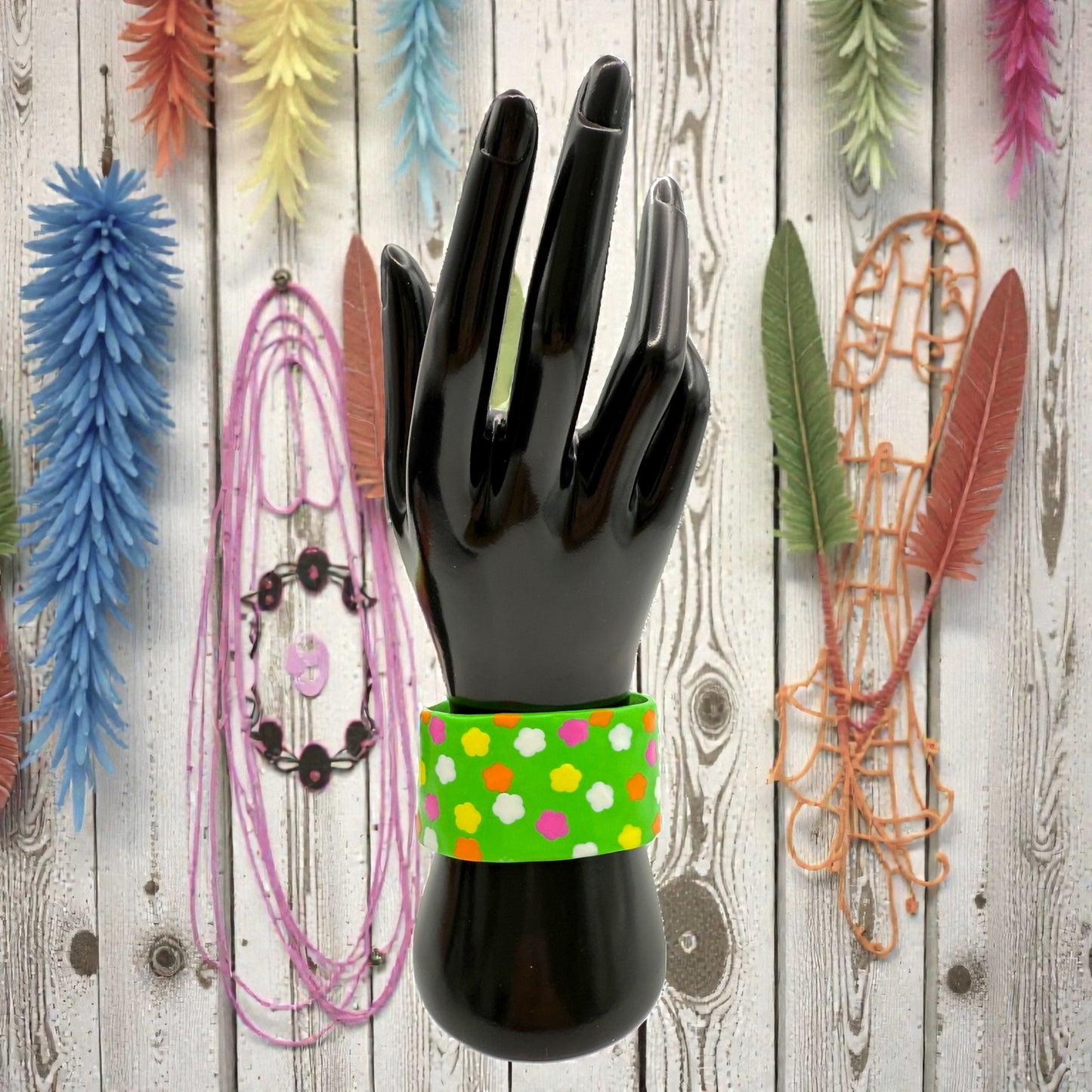 These Pretty Things Green Fluorescent Cuff on a black hand mannequin with coloured feathered leaves in the background