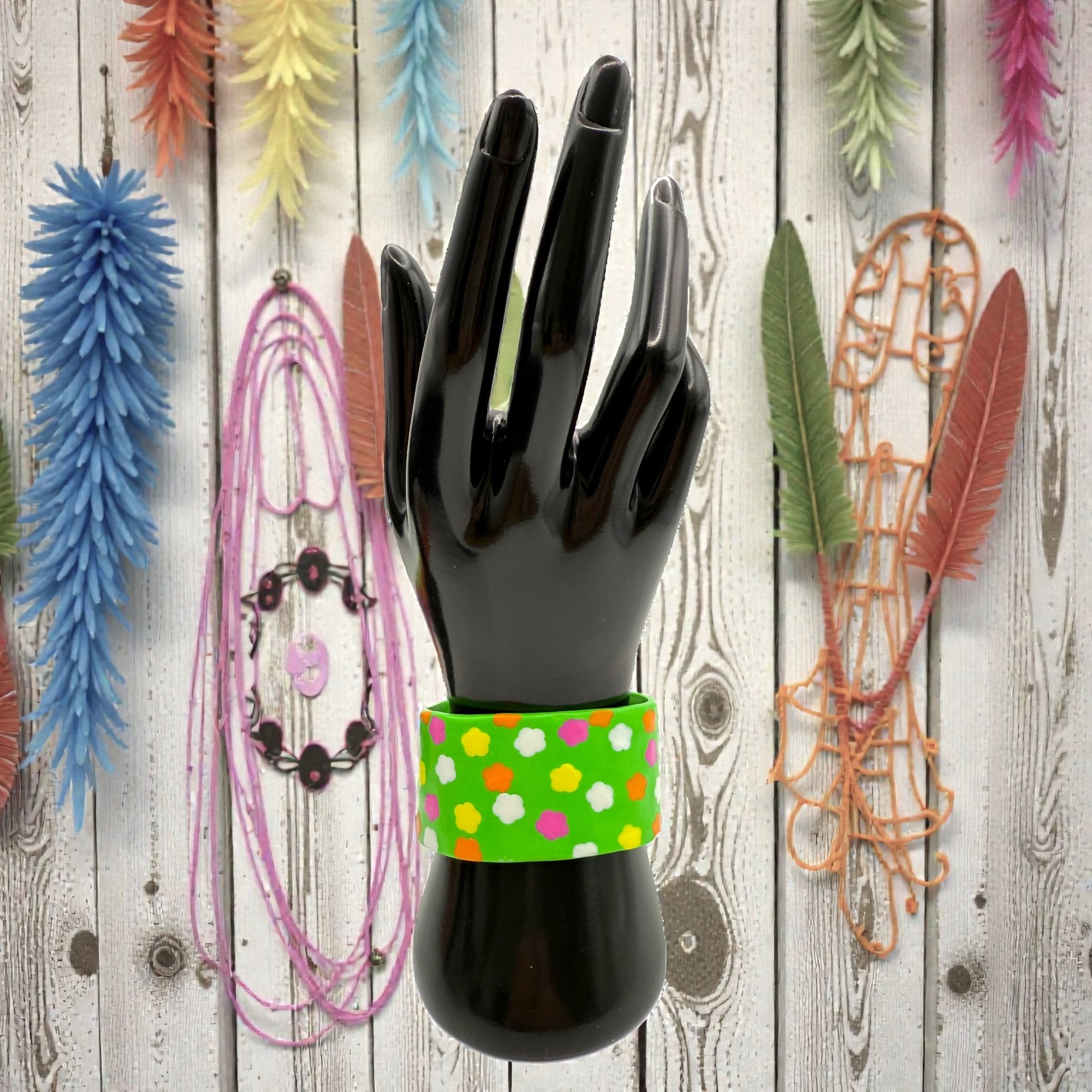 These Pretty Things Green Fluorescent Cuff on a black hand mannequin with coloured feathered leaves in the background