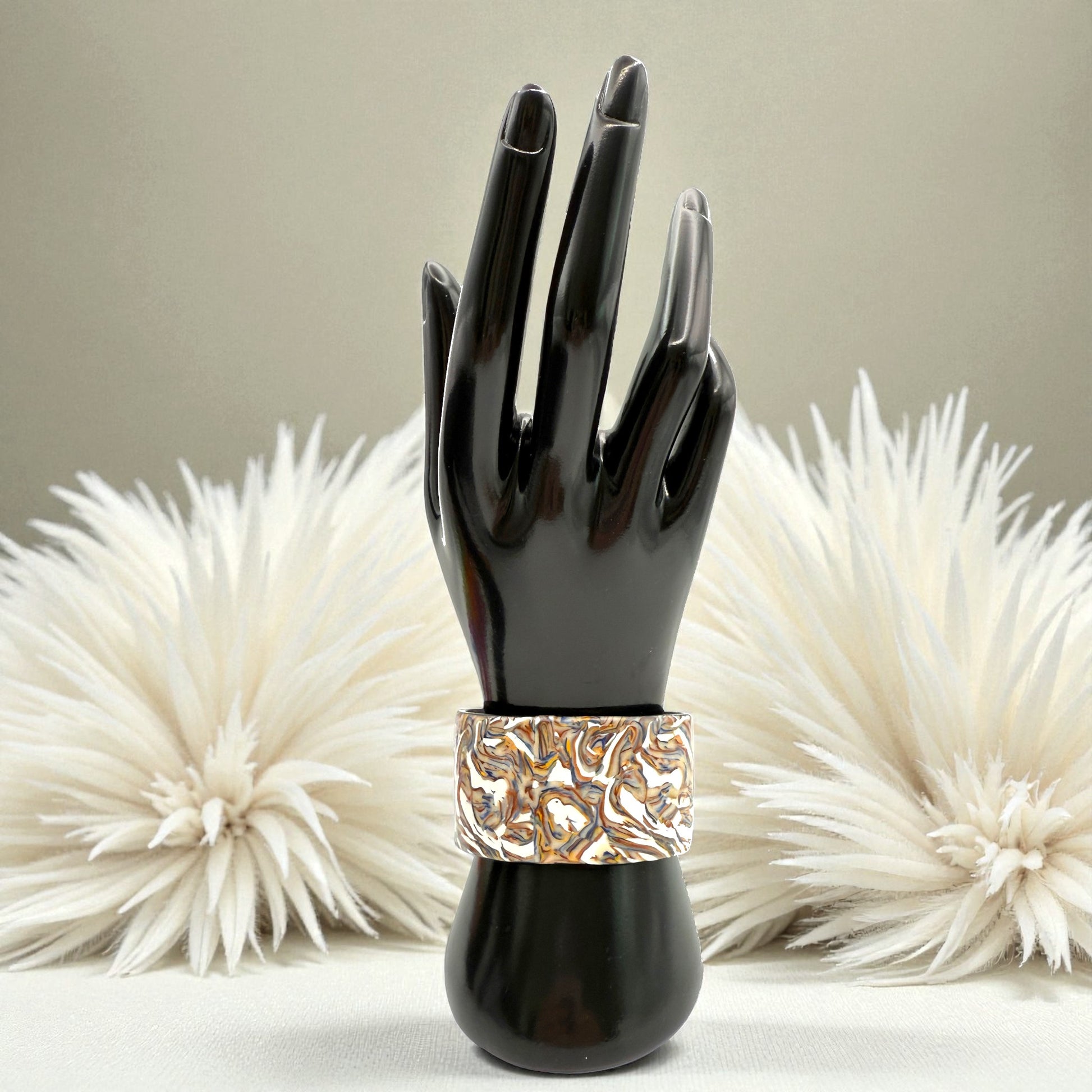 These Pretty Things Marbled Earth Cuff on a black wrist/hand demonstration bust set in front of a beige feather boa oon a beige colour table