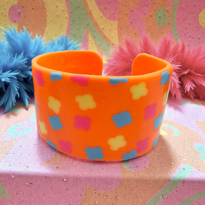 These Pretty Things Orange Fluorescent Cuff on a muted pink base with blue and yellow feather boas in the background