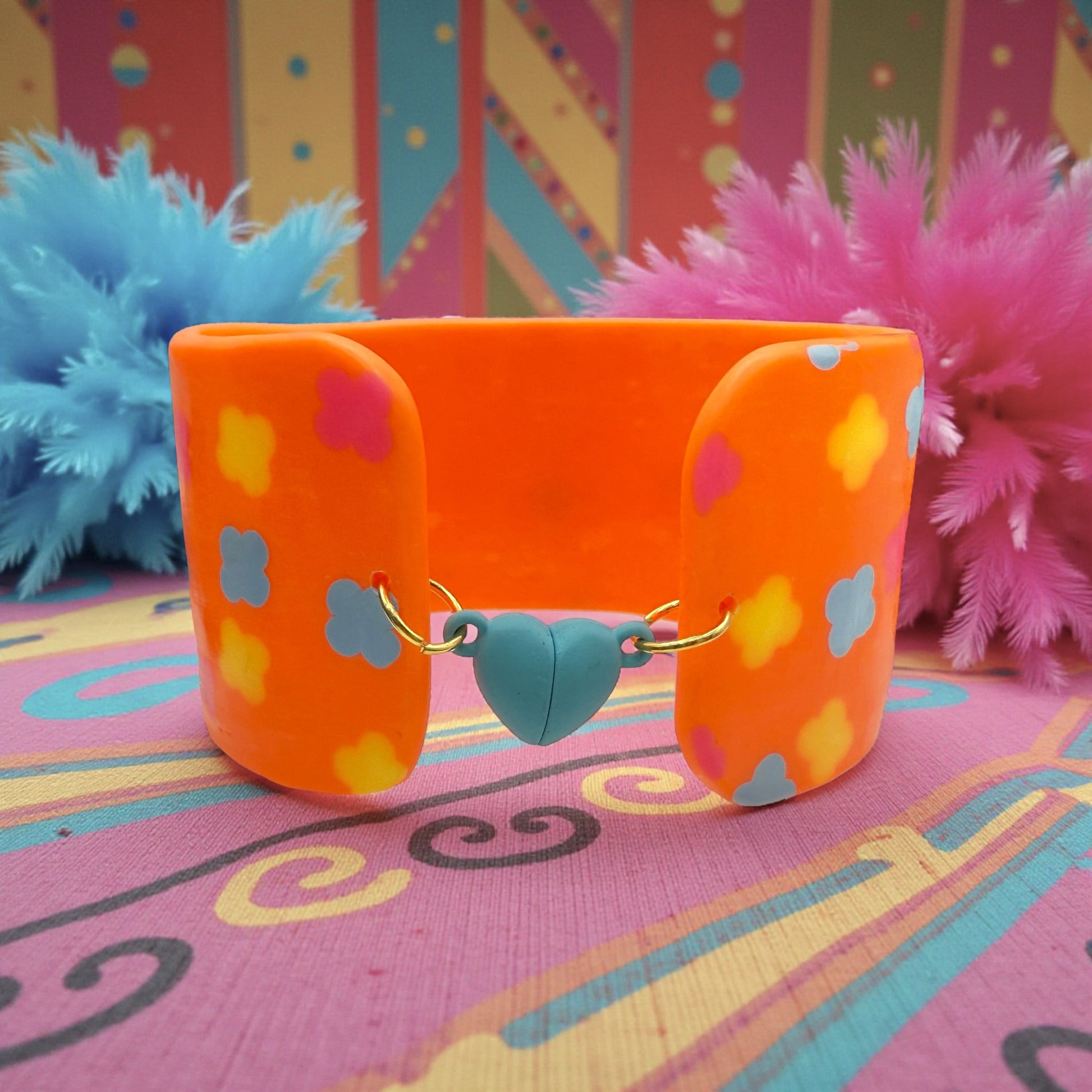 These Pretty Things Orange Fluorescent Cuff on a patterned pink base with blue and pink feather boas in the background