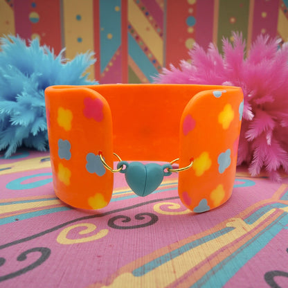 These Pretty Things Orange Fluorescent Cuff on a patterned pink base with blue and pink feather boas in the background