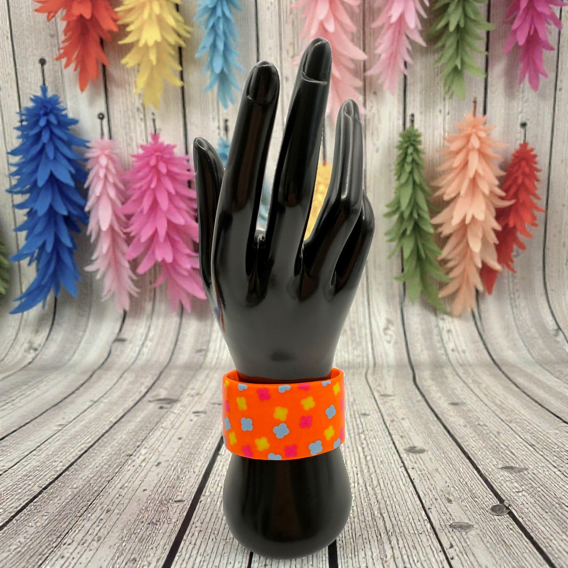 These Pretty Things Orange Fluorescent Cuff on a black hand mannequin with multiple coloured feather boas in the background