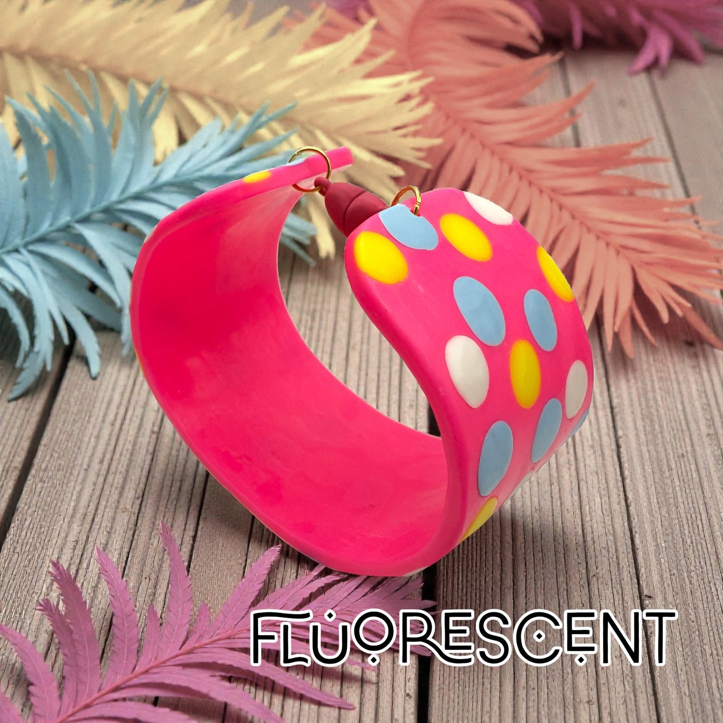 These Pretty Things Pink Fluorescent Cuff on a slated wood base with a variety of pale coloured feathers in the background
