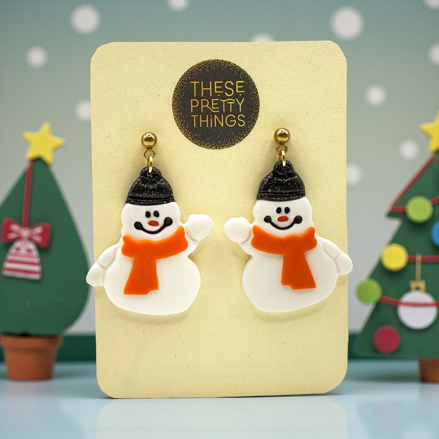 Snowmen earrings on a 'These Pretty Things' presentation card with Christmas trees in the background.