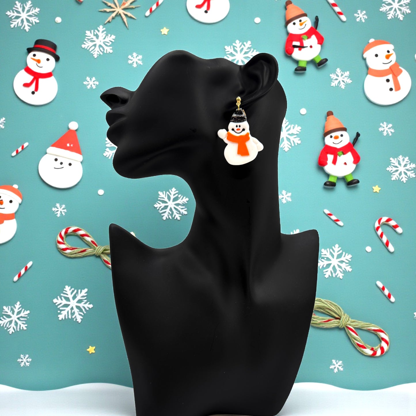 Snowman earring on a black mannequin against a Christmas-themed blue background with snowflakes, candy canes, and snowmen.